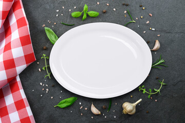 Classic Italian cooking template - top view of white blank ceramic plate on vintage black stone kitchen countertop with  red checkered tablecloth and selection of herbs and spices.