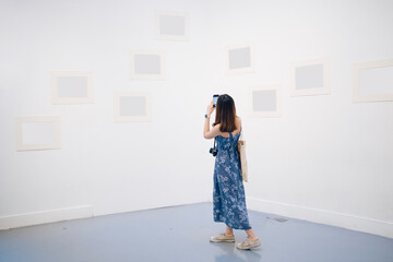 Young asian thai woman in blue dress taking picture with smartphone in white gallery.