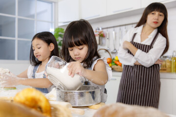 Two little cute Asian girls, 3 years and 7 years old, learning how to make bread and bakery with a curious and happy smiley face. Their mom standing and looking behind them