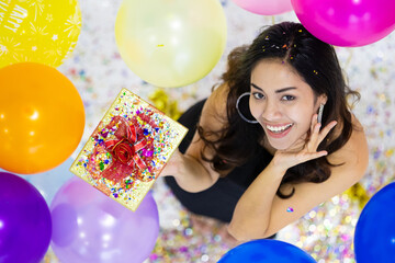 Two young and cute Asian women friends in black sexy dress holding gift boxes celebrate the new year festival with bright color balloons around them. Take from top-down angle