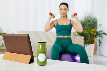 Glass bottle of green smoothie on table next to tablet computer and fit woman exercising with dumbbells in background