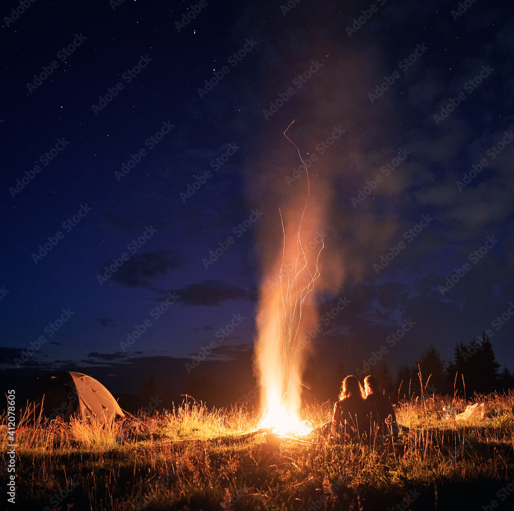 Beautiful view of night starry sky over grassy hill with camp tent, campfire and hikers. Young man and woman tourists having a rest near bonfire. Concept of hiking, night camping and relationship. - obrazy, fototapety, plakaty 