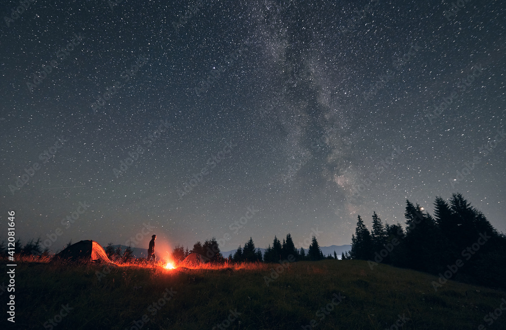 Male hiker standing near campfire and tent under beautiful night sky with stars. Magnificent view of blue starry sky and Milky way under grassy hill. Concept of travelling, hiking and night camping. - obrazy, fototapety, plakaty 