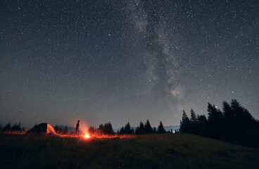 Wall Mural - male hiker standing near campfire and tent under beautiful night sky with stars. magnificent view of