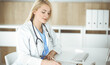 © Iryna - Woman-doctor at work while sitting at the desk in hospital or clinic. Blonde cheerful physician filling up medication history record form