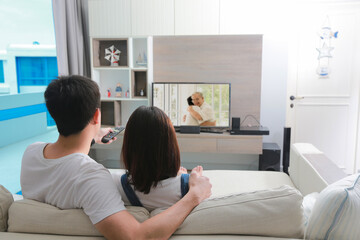 Back view of lovers hugging and sitting on sofa and watching TV in living room.