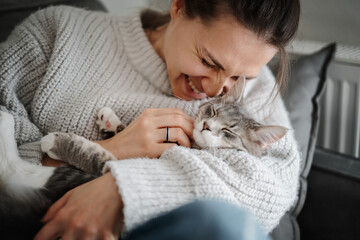 Beautiful cheerful young woman with a cute gray cat in her arms at home on the sofa, friendship and love for pets