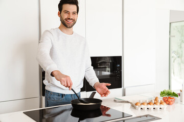 Happy handsome young man smiling while cooking scrambled eggs