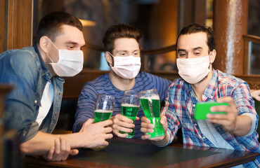 Wall Mural - friendship, health and st patrick's day concept - male friends wearing face protective medical mask for protection from virus disease drinking green beer and taking selfie with smartphone at bar