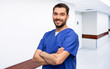 © Syda Productions - healthcare, profession and medicine concept - happy smiling doctor or male nurse in blue uniform with crossed arms over hospital corridor background