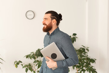 Portrait of young bearded man in office holding laptop.