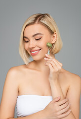 Wall Mural - Smiling woman in white top using jade roller isolated on grey