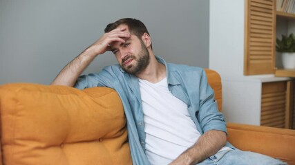 Poster - Sick ill tired sad young man in casual white t-shirt blue shirt sit on sofa couch put hands on head rub temples have headache migraine spend time living room at home in own house. Real estate concept