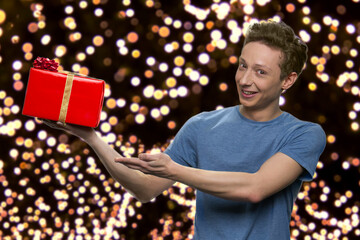 Guy in t-shirt is showing a red gift box. Thousands glowing lights on the background.