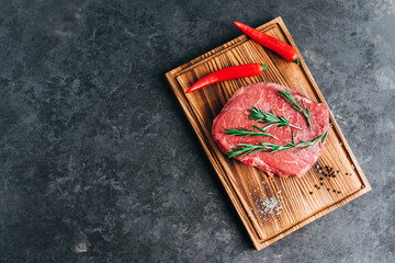 Wall Mural - Raw beef steak on wooden board and black background with rosemary, chili pepper and spices, copy space.