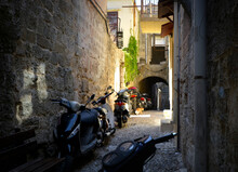 A Stray Alley Cat Wanders Down A Narrow Alley Filled With Motorcycles On The Mediterranean Island Of Rhodes Greece