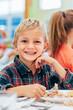 © MyMicrostock/Stocksy - Cute blond boy looking at camera while eating in classroom