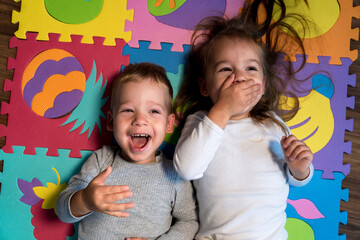 childhood, family friendship, games - close up portrait Two funny joy happy smiling little toddler peschool kids siblings twins brother with sister have fun lie laughing on puzzles mat at home indoors