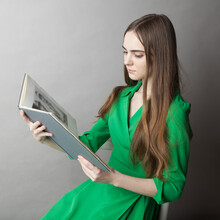Serious Romantic Teenager Girl With Long Hair In Green Dress Reading A Book While Sitting Indoors With Gray Walls
