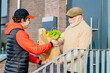 © Iryna - Profile shot of an elderly senior man taking to a food delivery male with a grocery box outdoor with building on background. Meal basket as social help and support.