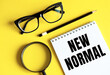 © Nastassia - NEW NORMAL text written on a notebook with glasses, magnifying glass and pencil.