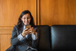 A young businesswoman or lawyer sitting on a sofa in the office looking at a mobile phone device and smiling.