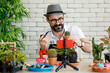 © Bangkok Click Studio - An internet seller, a beautiful beard man wearing hat and eyeglasses sitting in home indoor garden make an online presenting and live broadcast for sale his cactus in home garden