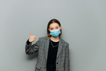 Serious woman in formalwear and protective mask stands on a background of gray wall and shows thumbs up, looking at the camera with a serious face.