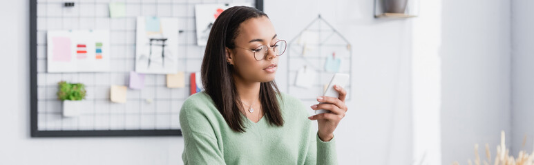 Canvas Print - young african american architect in eyeglasses messaging on smartphone, banner