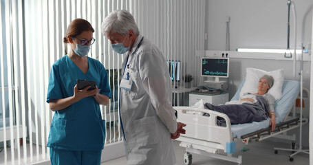 Doctor and nurse in safety mask discussing diagnosis on tablet with woman patient lying in bed