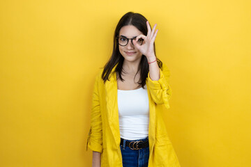 Wall Mural - Young brunette businesswoman wearing yellow blazer over yellow background doing ok gesture shocked with smiling face, eye looking through fingers