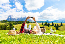 Young couple taking a selfie holding smart phone mobile lying on the grass at park - Tourist at holiday visiting Siusi alps, Italy - Bright filter