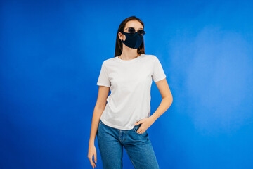 Wall Mural - Stylish girl in glasses and mask wearing white t-shirt posing in studio on blue background