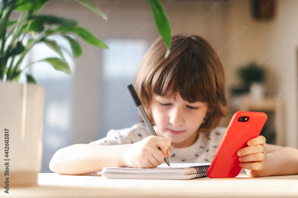 Back to school. Distance learning online education. Caucasian smile kid boy studying at home with mobile smart phone and doing school homework. Thinking child siting at table with cell phone