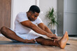Portrait of flexible African-American man practicing yoga sitting in Janu Sirsasana exercise, Head to Knee Forward Bend pose, on floor during working out at bright living room.