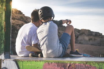 Back view of teens talking about tricks and jumps with skateboard. Young boys sitting on half pipe ramp resting after having fun with boards, looking away. Youth togetherness, sport, positive concept