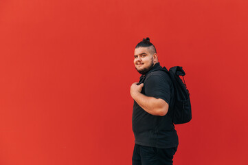positive overweight young man in stylish casual clothes stands with a backpack on his back against a