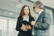 © Mangostar - Senior executive manager discussing with employee, holding tablet and standing in conference room. Red-haired businesswoman looking at businessman and listening him. Business and management concept