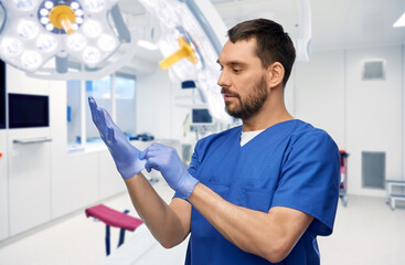 Sticker - medicine, healthcare and surgery concept - doctor or male nurse in blue uniform putting medical gloves on over operating room at hospital background