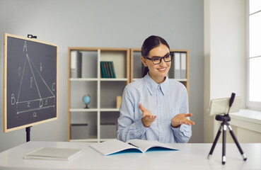 Smiling school math teacher or private tutor giving engaging virtual lesson, using cell phone on tripod. Happy young woman in glasses explaining geometry problem in remote online class via video call