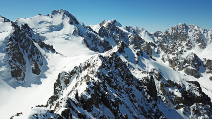 Snow-capped mountains and blue sky. View from a drone. Huge peaks covered with snow, steep cliffs. Blue, clear sky above the mountains. Flying over the peaks. Below you can see green trees and tarva.