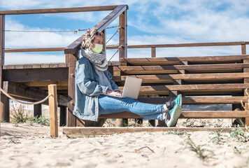 Canvas Print - Young Caucasian female student with a face mask working on the computer on a boardwalk on a beach