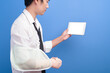 © tonefotografia - Young businessman with an injured arm in a sling using a tablet over blue background in studio, insurance and healthcare concept