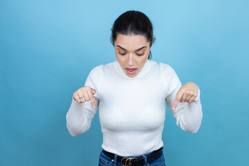 Wall Mural - Young caucasian woman wearing white sweater over blue background surprised, looking down and pointing down with fingers and raised arms