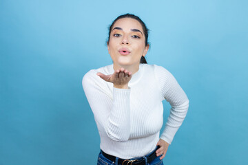 Wall Mural - Young caucasian woman wearing white sweater over blue background looking at the camera blowing a kiss with hand on air being lovely and sexy. Love expression.