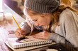 © Mint Images - Teenage girl in a woolly hat drawing with a pencil on a sketchpad.