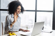 © Face Stock - Mixed-race businesswoman in casual clothes with Afro hairstyle sitting in the office with panoramic window, studying or working online, having a virtual meeting, saying hello, waving at laptop screen