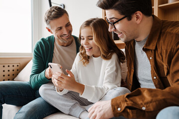 Wall Mural - Happy gay family smiling and using mobile phone while sitting on couch