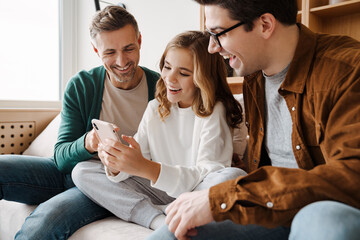 Wall Mural - Happy gay family smiling and using mobile phone while sitting on couch