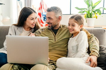 Wall Mural - Happy military man smiling and hugging his family while using laptop